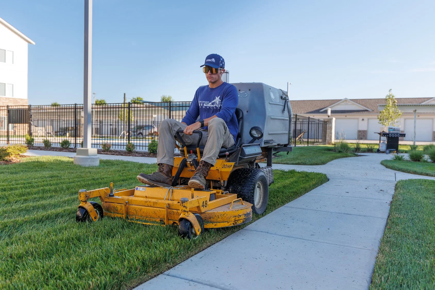 Operator on yellow zero-turn mower cutting residential lawn with concrete pathway and homes visible