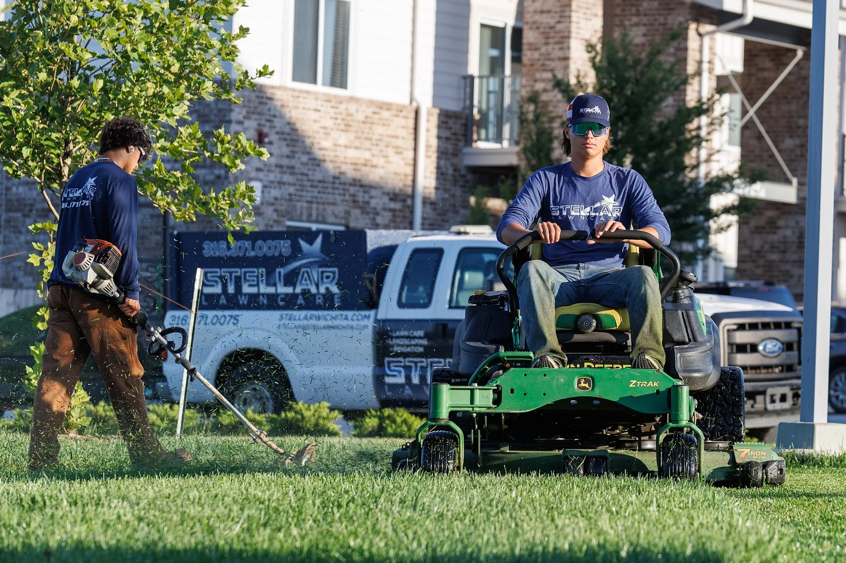 Crew operating green zero-turn mower and using string trimmer on residential lawn with brick home